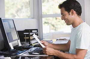 Man in home office using computer holding paperwork and smiling