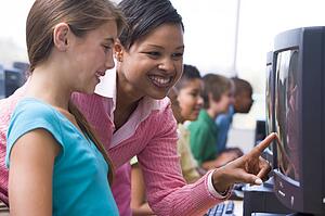 Teacher helping student at computer terminal with students in background (depth of field/high key)