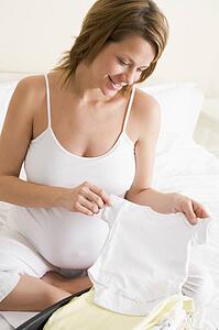 Pregnant woman packing baby clothing in suitcase smiling