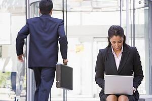 Businesswoman outdoors in front of building using laptop with businessman in background (high key/blur)
