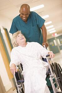An Orderly Pushing A Senior Woman In A Wheelchair Down A Hospital Corridor