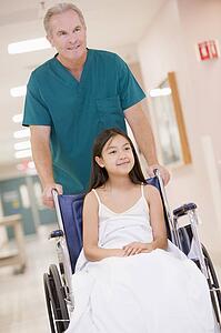 An Orderly Pushing A Little Girl In A Wheelchair Down A Hospital Corridor