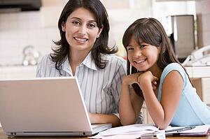 Woman and young girl in kitchen with laptop and paperwork smiling