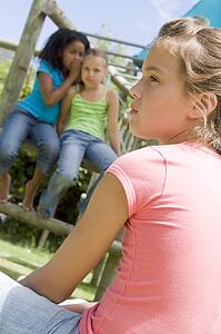 Two young girl friends at a playground whispering about other girl in foreground