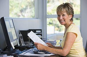Woman in home office with computer and paperwork smiling