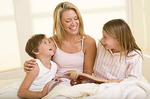 Woman with two young children sitting in bed reading book and smiling