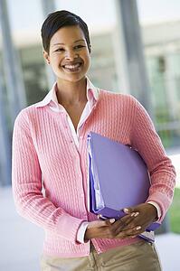 Teacher standing outside school holding binders and smiling (selective focus)