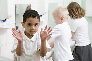 Students in bathroom at sinks washing hands with one holding up soapy hands (selective focus)