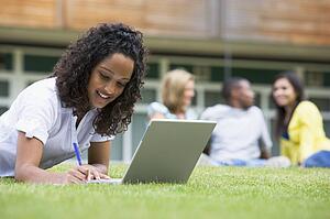 Student outdoors on lawn using laptop with other students in background (selective focus)