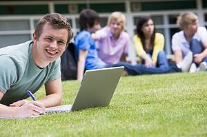Student outdoors on lawn using laptop with other students in background (selective focus)