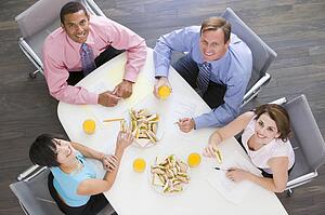 Four businesspeople at boardroom table with sandwiches smiling