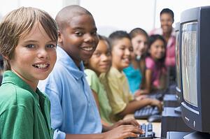 Six children at computer terminals with teacher in background (depth of field/high key)