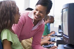 Teacher helping a student at computer terminal with students in background (selective focus/high key)