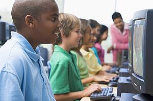 Six children at computer terminals with teacher in background (depth of field/high key)