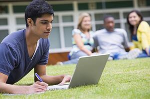 Student outdoors on lawn using laptop with other students in background (selective focus)