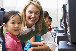 Teacher helping student at computer terminal with students in background (depth of field/high key)