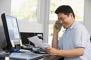 Man in home office with computer and paperwork on telephone smiling