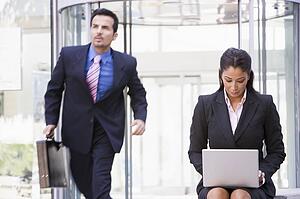Businesswoman outdoors in front of building using laptop with businessman in background (high key/blur)