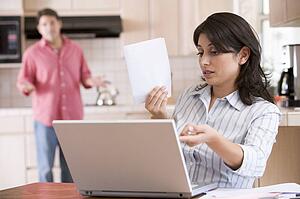 Woman in kitchen with paperwork using laptop with man in background