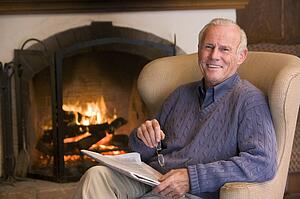Man sitting in living room by fireplace with newspaper smiling