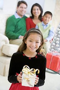 Young Girl Standing Holding Christmas Present, With Her Parents And Brother Sitting In The Background