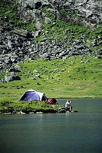 Camper outdoors at campsite by lake drinking beverage (far away)