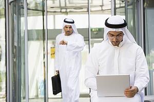 Businessman outdoors using laptop with businessman walking in background checking watch (selective focus)