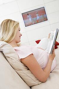 Pregnant woman reading magazine with television in background smiling