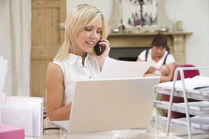 Woman in home office with laptop and telephone with mother and baby in background