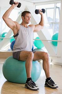Man Using Hand Weights On Swiss Ball At Gym