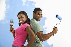 Couple next to half-painted wall as they stand back to back with paint utensils