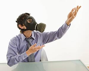 Businessman sitting in white room wearing gas mask gesturing to unknown person