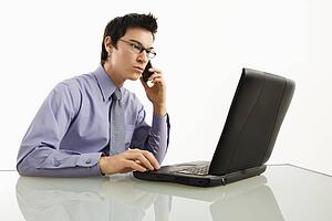 Businessman sitting at desk working on laptop talking on cellphone