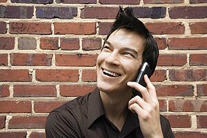 Smiling young Asian man next to brick wall talking on cell phone.