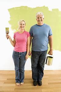 Portrait of happy adult couple standing in front of half-painted wall with paint supplies 'American Gothic' style.