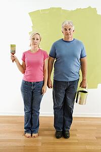 Portrait of unhappy adult couple standing in front of half-painted wall with paint supplies 'American Gothic' style