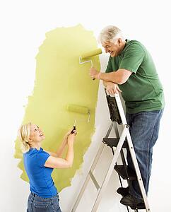 Middle-aged couple painting wall green with male on ladder.