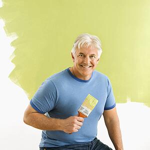 Smiling man sitting in front of half-painted wall with paintbrush
