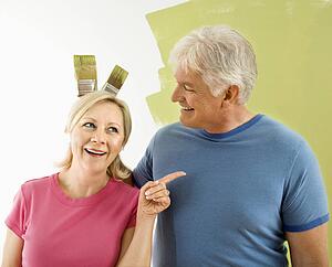 Portrait of happy adult couple standing in front of half-painted wall while man makes bunny ears with paintbrushes behind woman's head.