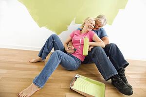 Portrait of happy adult couple sitting in front of half-painted wall with paint supplies laughing