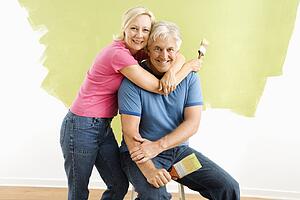 Portrait of smiling adult couple sitting in front of half-painted wall with paintbrushes