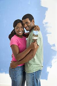 African American couple standing together hugging in front of half-painted wall and smiling at viewer.