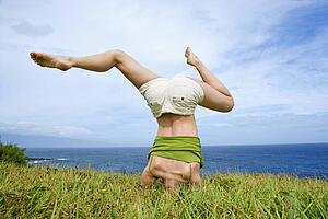 Woman doing headstand with legs askew in grass near ocean