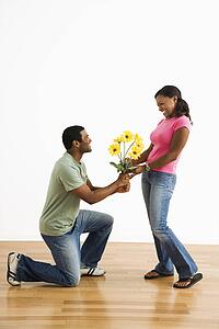 Male giving pretty female a bouquet of yellow flowers while on bended knee.