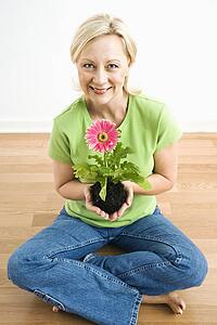 Portrait of smiling adult blonde woman sitting on floor holding pink gerbera daisy plant.