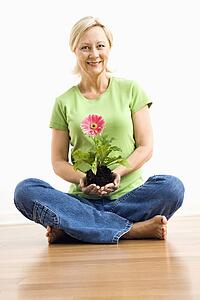 Portrait of smiling adult blonde woman sitting on floor holding pink gerbera daisy plant.