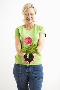 Portrait of smiling adult blonde woman standing holding pink gerbera daisy plant.