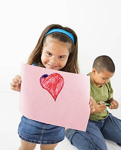 Young girl showing off drawing of heart while boy sits behind her