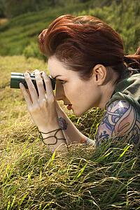 Attractive tattooed Caucasian woman lying on grass looking through binoculars 