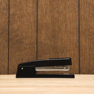 Black stapler on desk with wooden paneling.